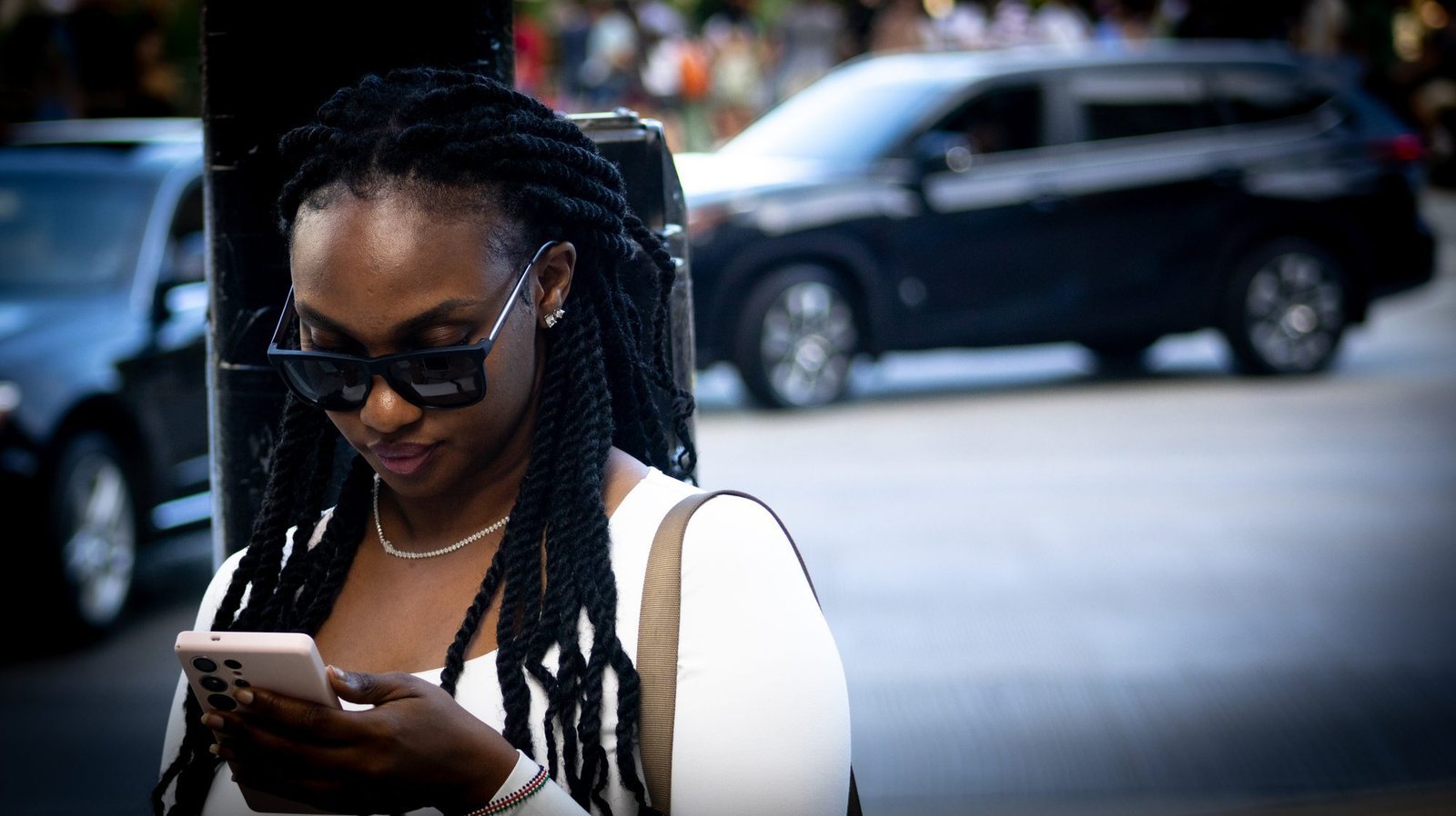 Woman with long twists and sunglasses looking down at her phone, a dark SUV blurred behind her